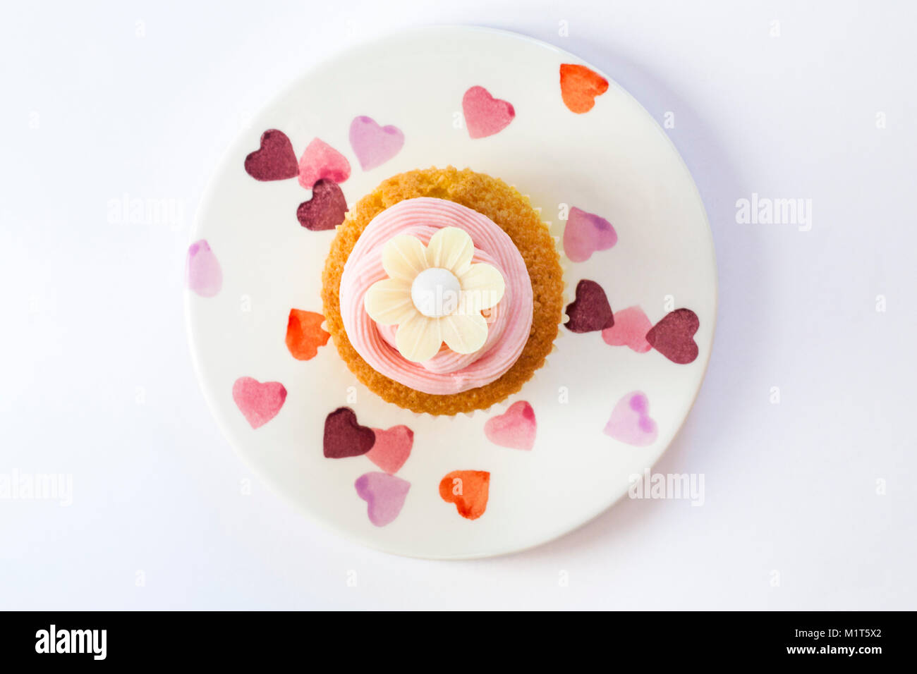 home made cupcake cake with strawberry pink frosting icing and rice paper edible daisy flower, on plate with hearts on isolated on white background Stock Photo