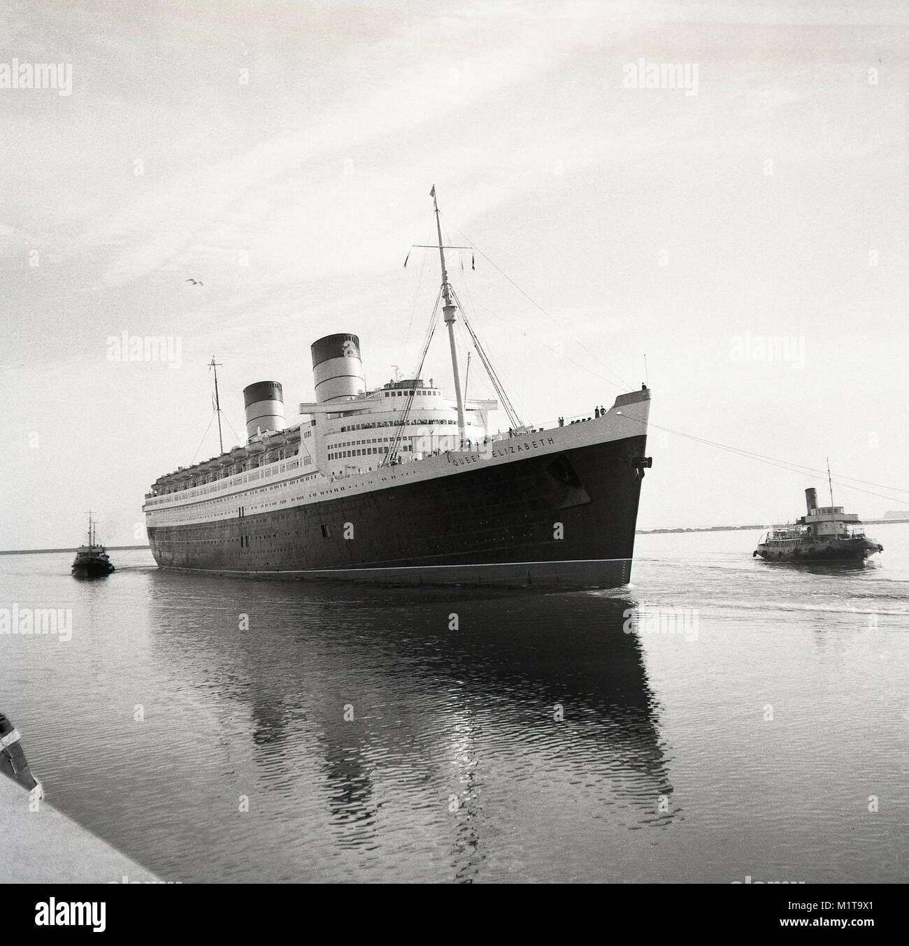 1950s, historical, tugboats pull the RMS Queen Elizabeth ocean liner into the calm waters of Southampton docks, England, UK. Named after the British Queen Mother, it was the largest passenger liner ever built at that time (1938) and remained so for 56 years thereafter. She was used as a troopship during WW2 and only became an ocean liner, as intended, in 1946. Stock Photo