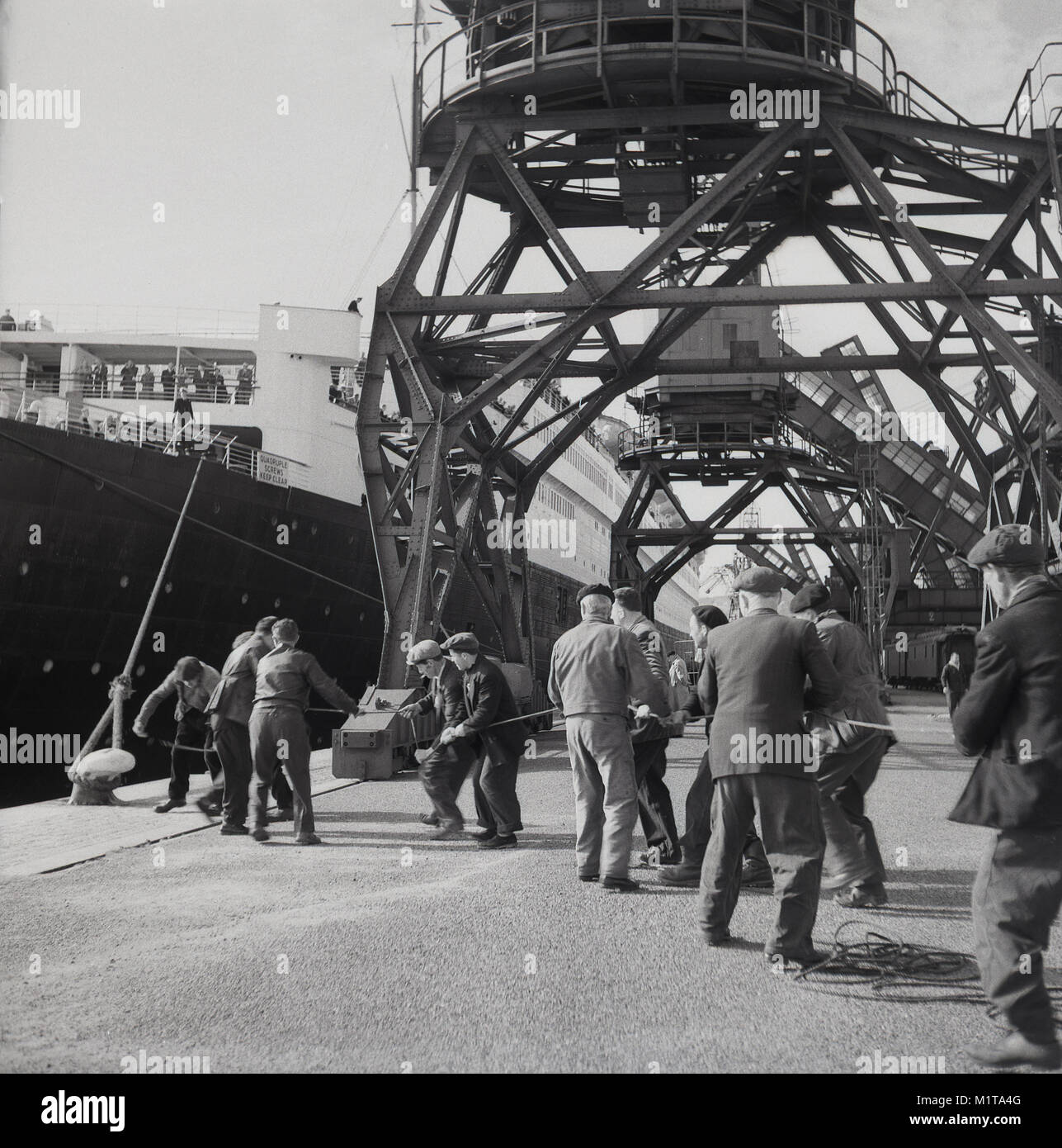1950s, historical, male dockers pulling a rope to tie the RMS Queen Elizabeth ocean liner in at Southampton docks, England, UK. Named after the British Queen Mother, it was the largest passenger ship ever built at that time (1938) and remained so for 56 years thereafter. She was used as a troopship during WW2 and only became an ocean liner as intended in 1946. Stock Photo