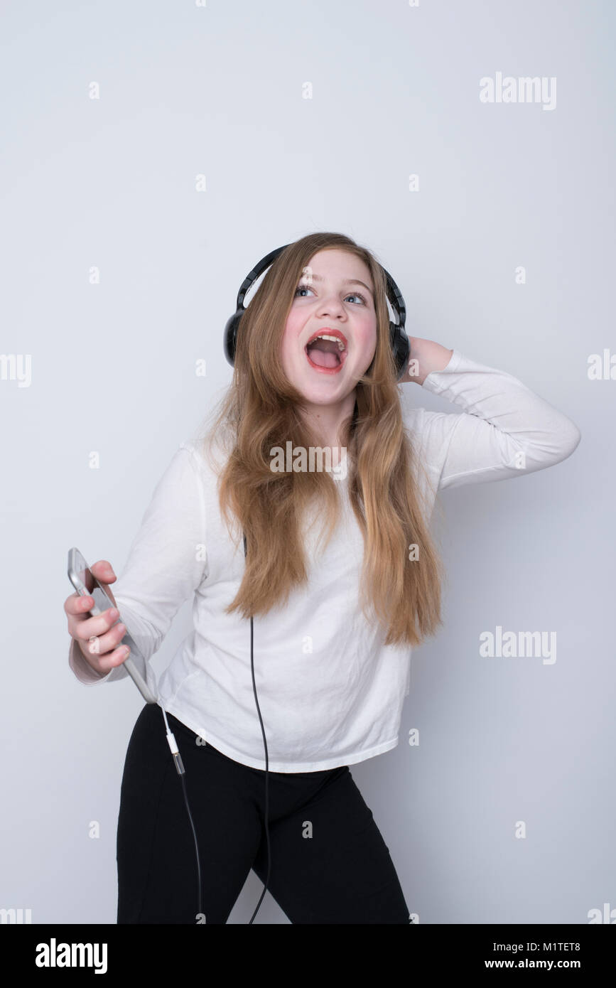 Smiling girl looking up while listening to music on smartphone with over-the-ear headphones and one hand behind head on white background Stock Photo