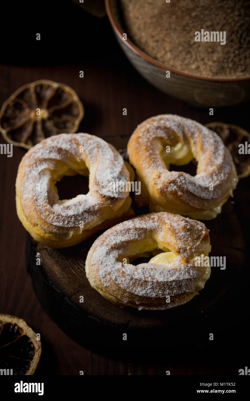 Vertical photo with three homemade puffs stuffed by sweet cream. The puffs are covered by powder sugar. Few dried orange rings are spilled around on d Stock Photo