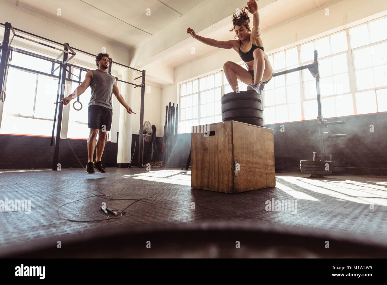 Fit young woman box jumping with man exercising with skipping ropes at a cross training style gym. Young couple training hard at the gym. Stock Photo