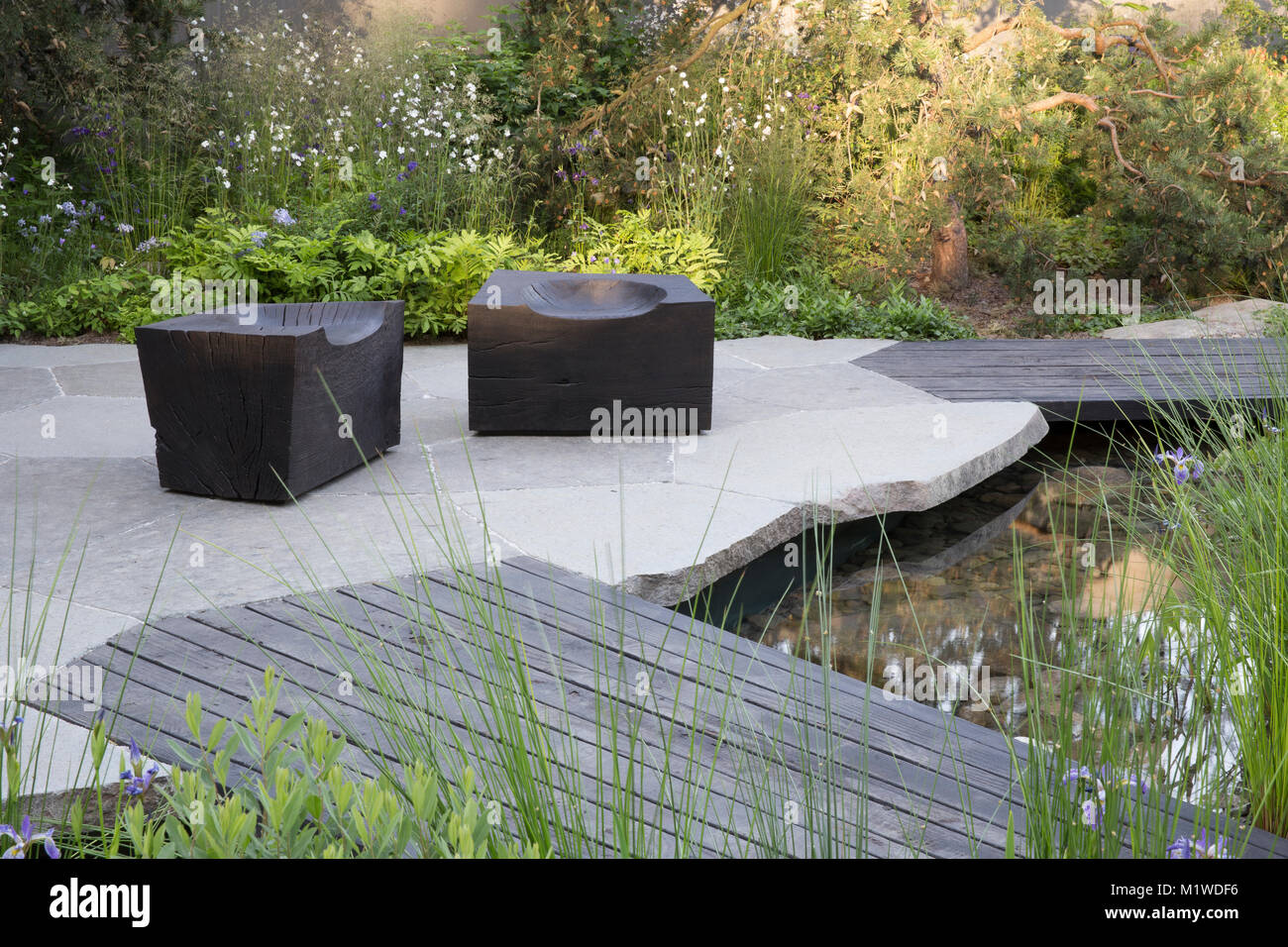 small modern garden space gardening with stone paved paving patio with wooden deck decking over a small pond pool  charred wooden cube seat UK Stock Photo