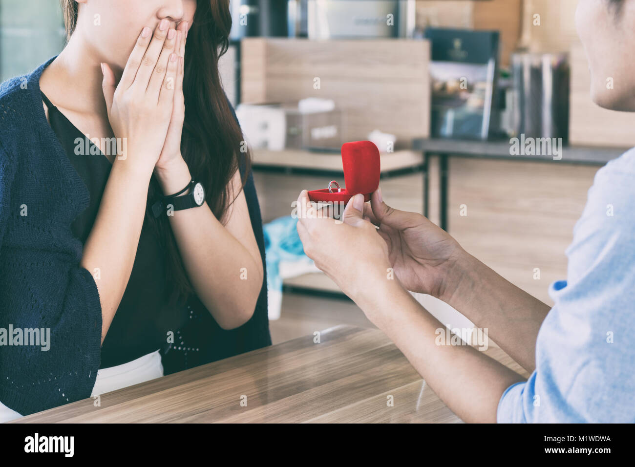 Asian man showing an engagement ring diamond to his amazed girlfriend in a restaurant with a warmth sunset outdoor in the background. Proposal concept Stock Photo