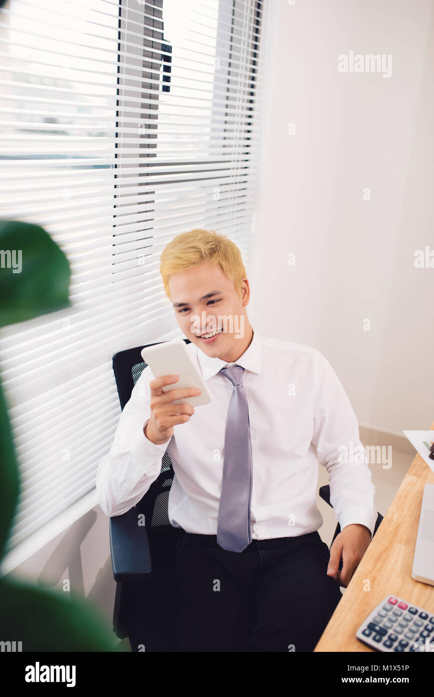 Texting to colleague. Confident young man in smart casual wear holding smart phone and looking at it while sitting at his working place in office Stock Photo