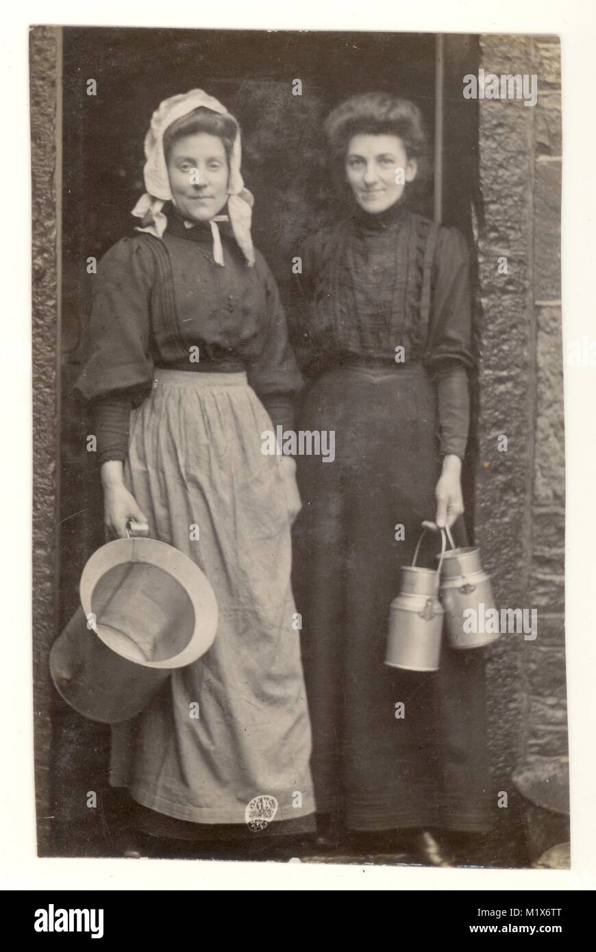 Original charming Edwardian era postcard portrait photograph of milk maid holding a milk pail and wearing a bonnet on the doorstep of a house with a lady customer, borough of Rosendale, Rawtenstall, Ramsbottom area, U.K. circa 1906 Stock Photo