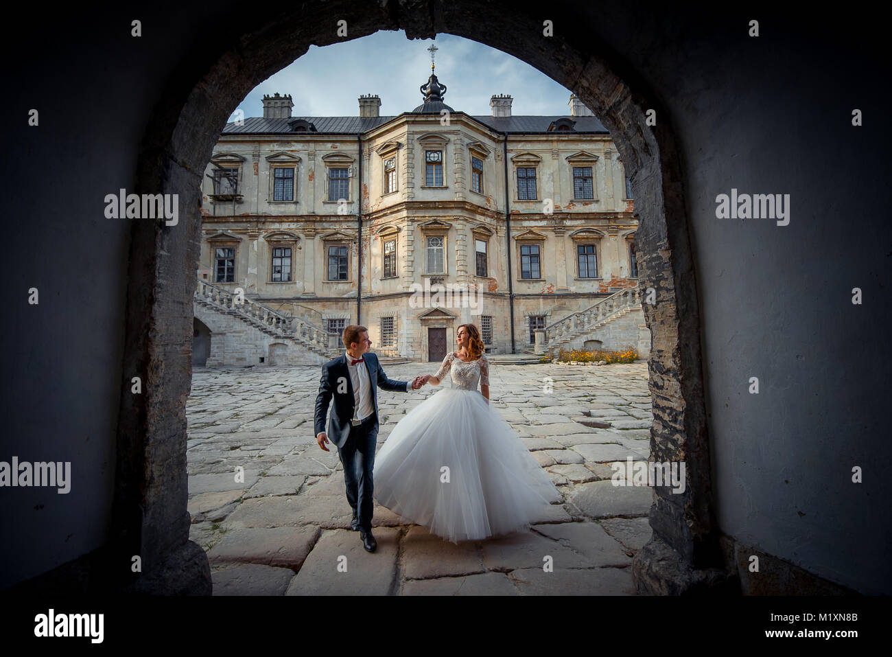Walking newlyweds at the background of the old castle. The groom is leading his attractive ginger hair bride by the hand. Look through the archway. Stock Photo