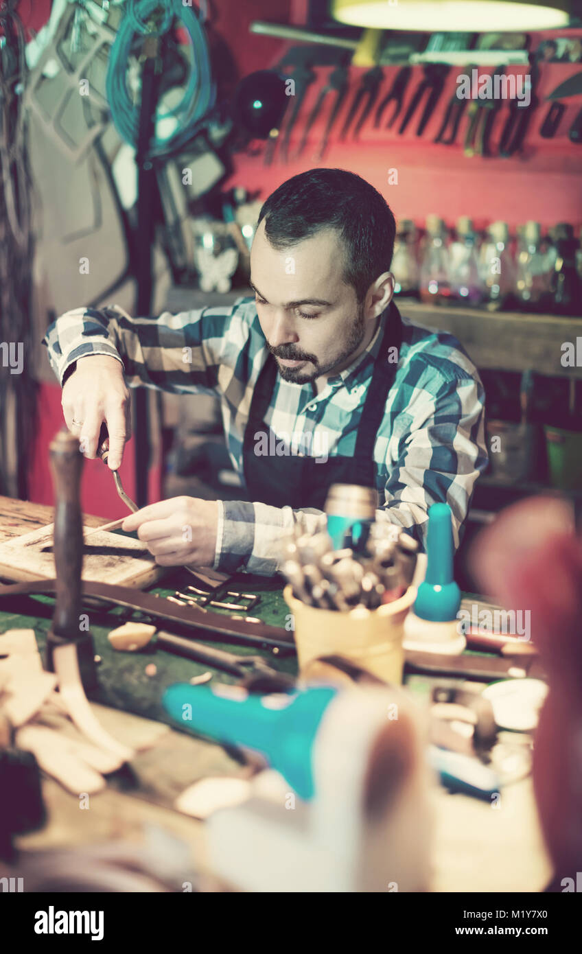 Young diligent friendly male worker working on leather for belt in