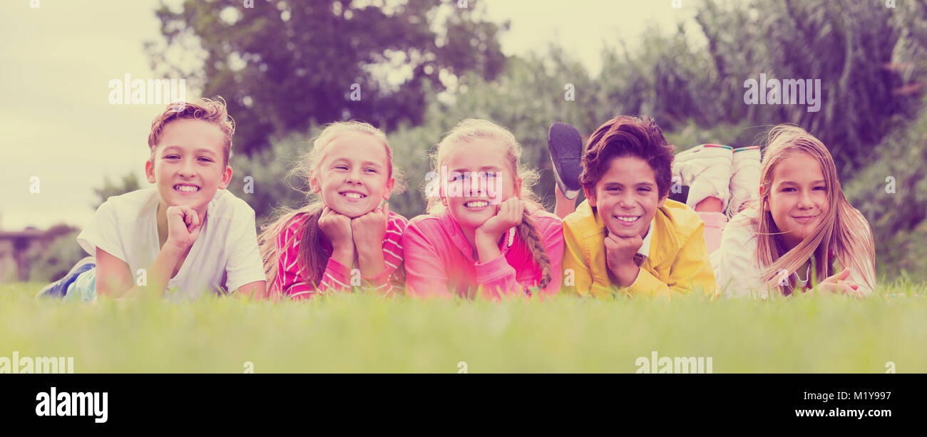 Company of five smiling kids are posing lying on grass in the park ...