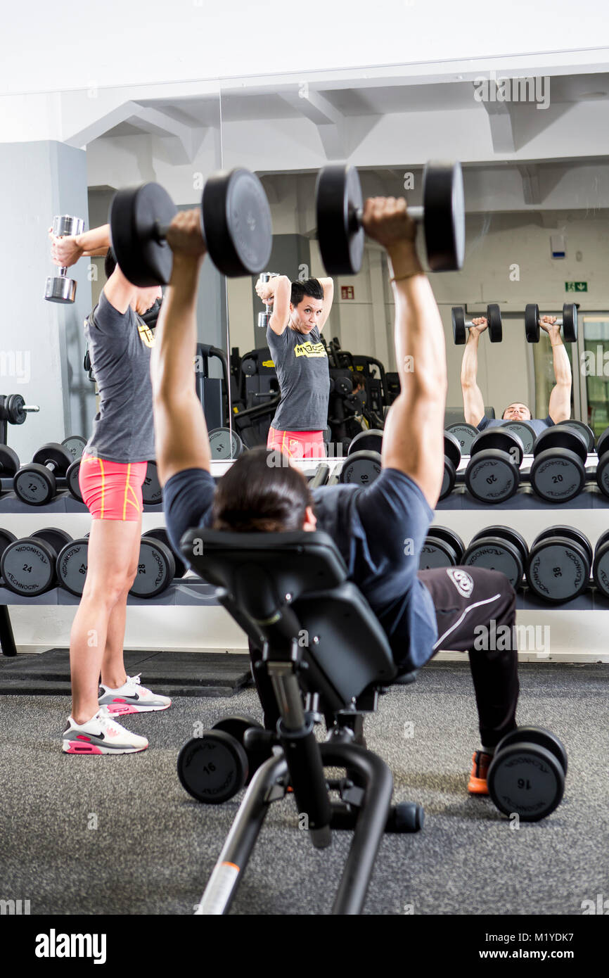 Man and woman training with dumbbells at the gym in front of a mirror. Woman is standing up and the man is sitting down. Focus is in the mirror. Stock Photo