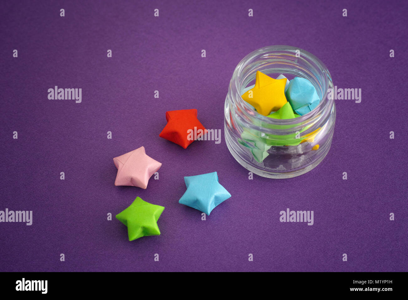 Colorful origami lucky stars spilling out of a jar. Purple background. Stock Photo