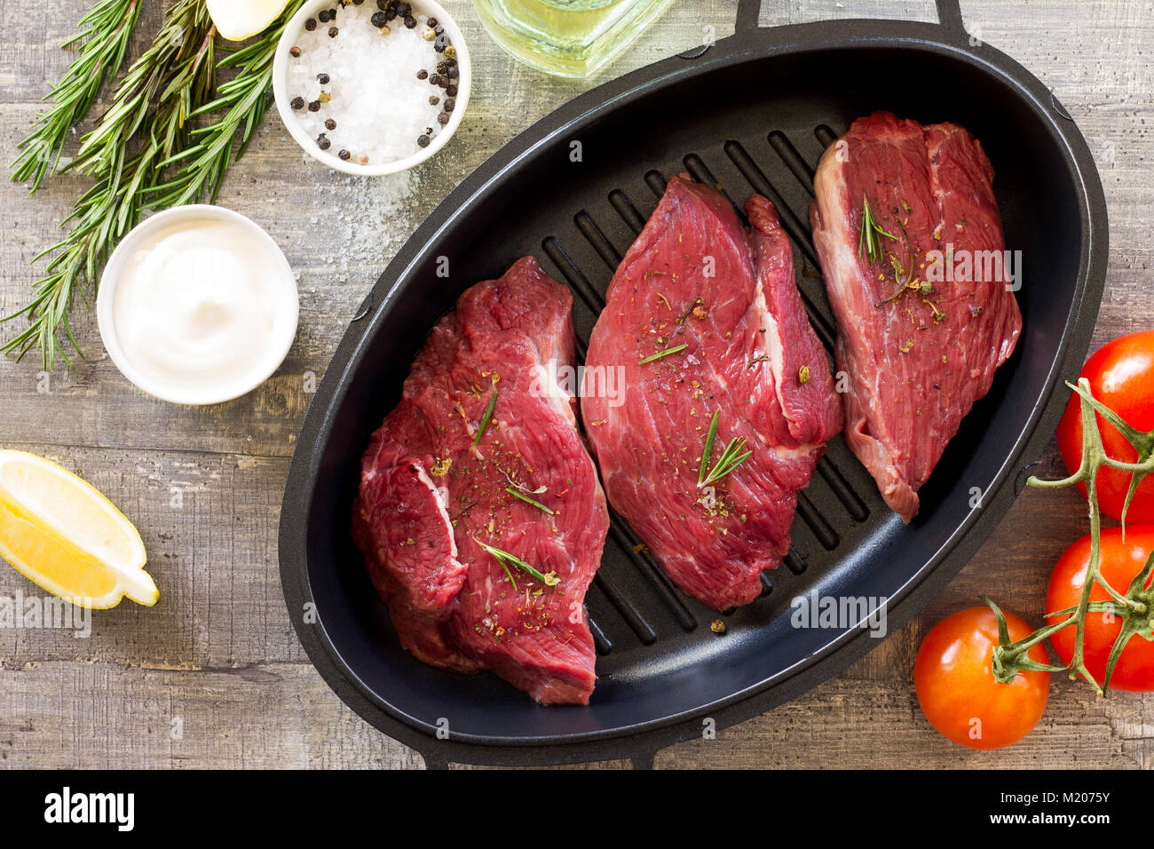 Fresh meat. Raw steak beef on a cast-iron grill frying pan, tomatoes, olive oil, spices and fresh rosemary on the kitchen table. Stock Photo
