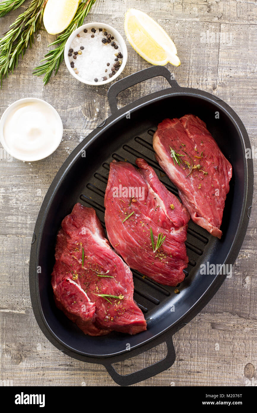 Fresh meat. Raw steak beef on a cast-iron grill frying pan, tomatoes, olive oil, spices and fresh rosemary on the kitchen table. Stock Photo