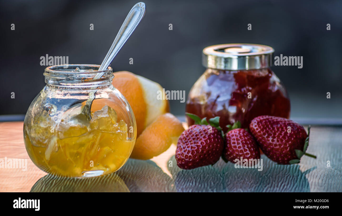 Colorful jams in glass jars and organic fresh fruit, closeup, macro food. Side view of yellow orange marmalade, fresh orange, red strawberry jam. Stock Photo