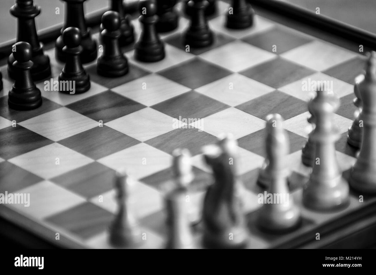 Cropped view of woodgrain chessboard with pieces blurred in starting positions, arranged diagonally and viewed at oblique angle, monochrome image Stock Photo