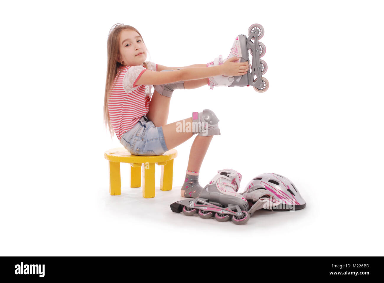 Charming broun-haired girl of school age in short jeans shorts and a pink t-shirt sitting on the chair and tries to foot roller skates. Isolated on wh Stock Photo