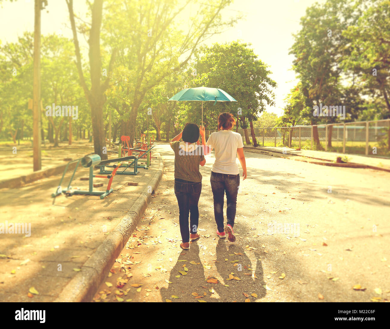 Best female friends sharing an umbrella while walking together in a park with strong sunshine during day time Stock Photo