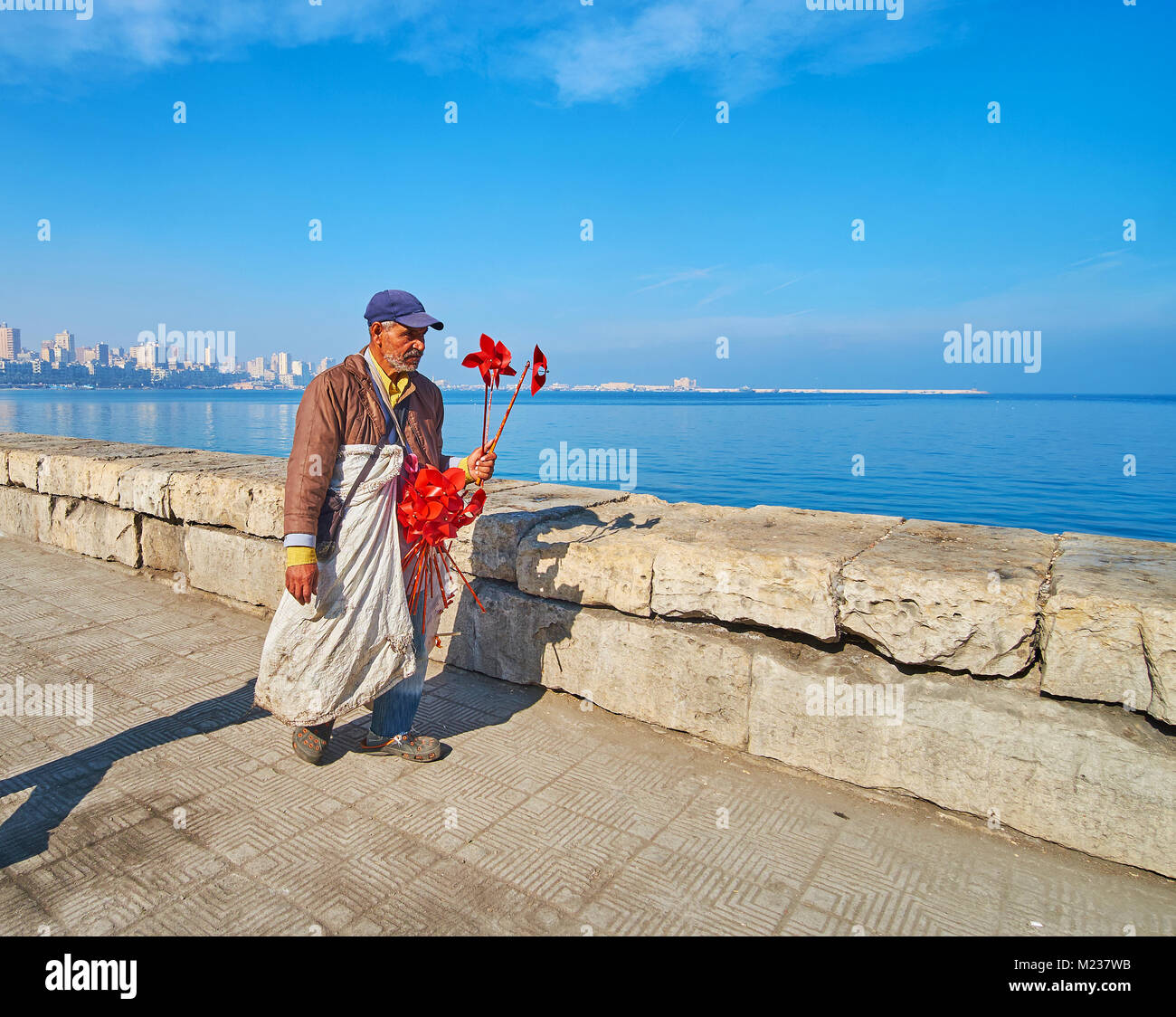 ALEXANDRIA, EGYPT - DECEMBER 17, 2017: The elderly vendor with red flower windmills goes along the Corniche promenade, on December 17 in Alexandria. Stock Photo