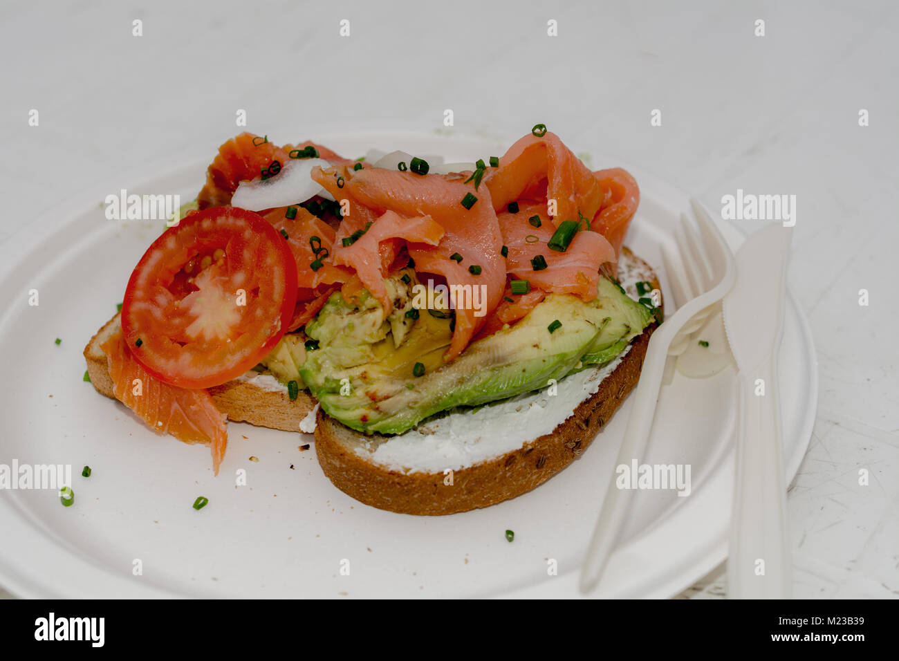 Gourmet Mexican avocado toast garnished with cream cheese, lemon zest, smoked salmon, and tomato, on white plate and table served with plastic fork an Stock Photo