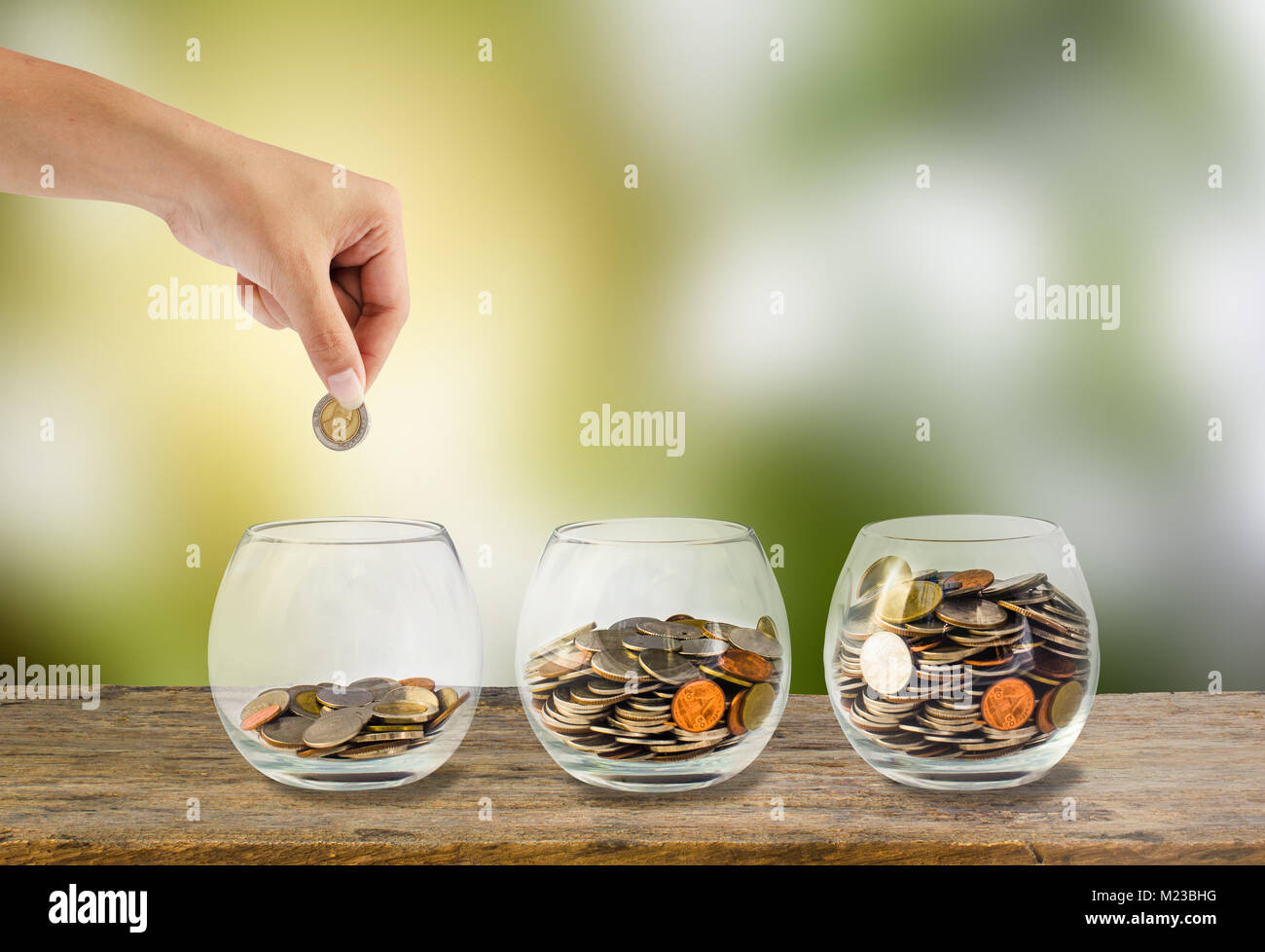 Saving money concept. Hand holding putting coins into three step clear glass bottle on wooden table with green blurred background and light. Conceptua Stock Photo