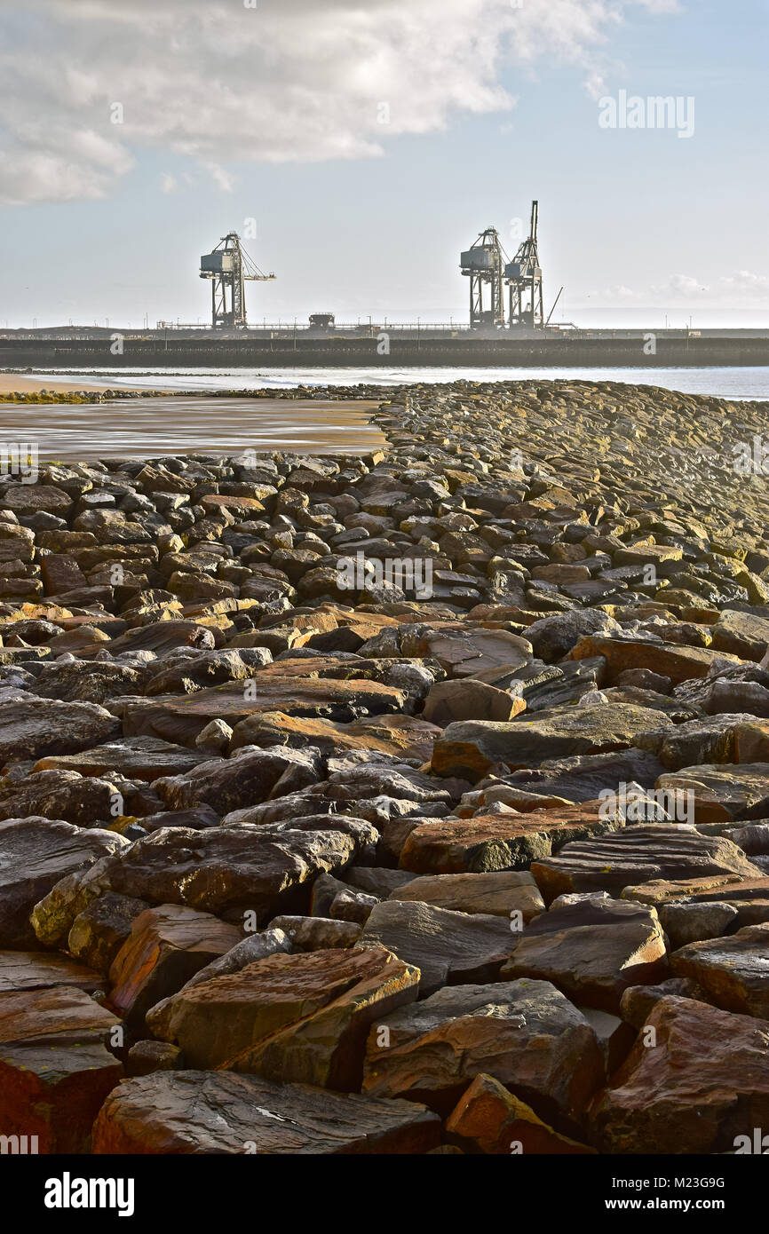 Heavy stones forming part of the breakwater at the beach in Aberavon with three large cranes of Port Talbot steelworks docks in background. Stock Photo
