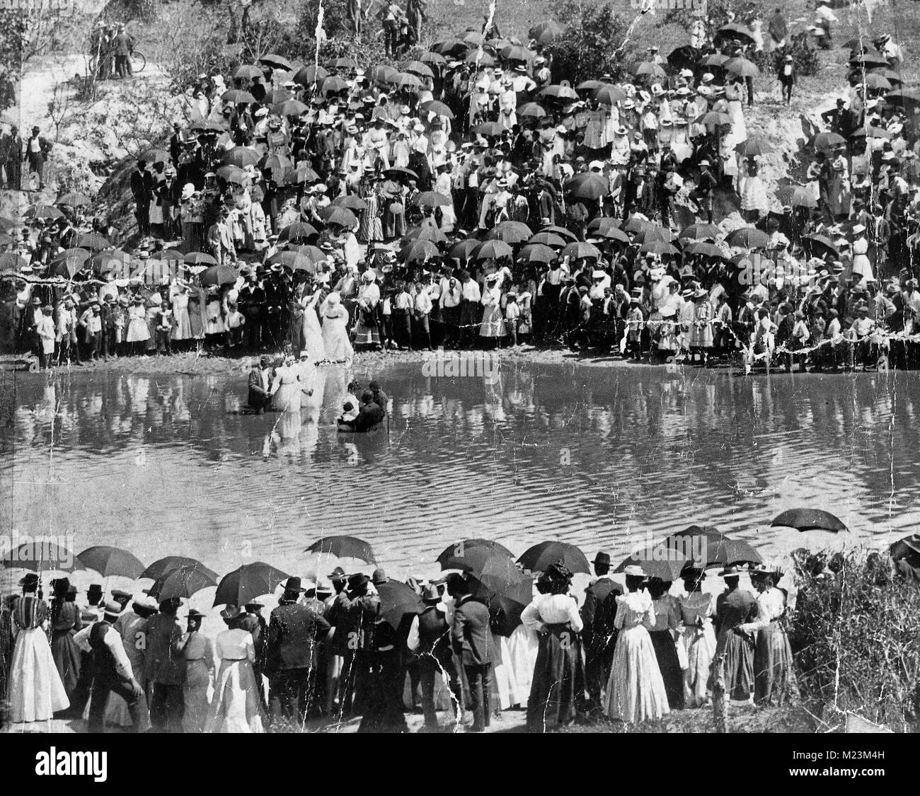 A large group of African-American spectators stands on the banks of Buffalo Bayou to witness a baptism. Many umbrellas are present indicating an effort to provide some shade from the heat of the day, circa 1900 Stock Photo