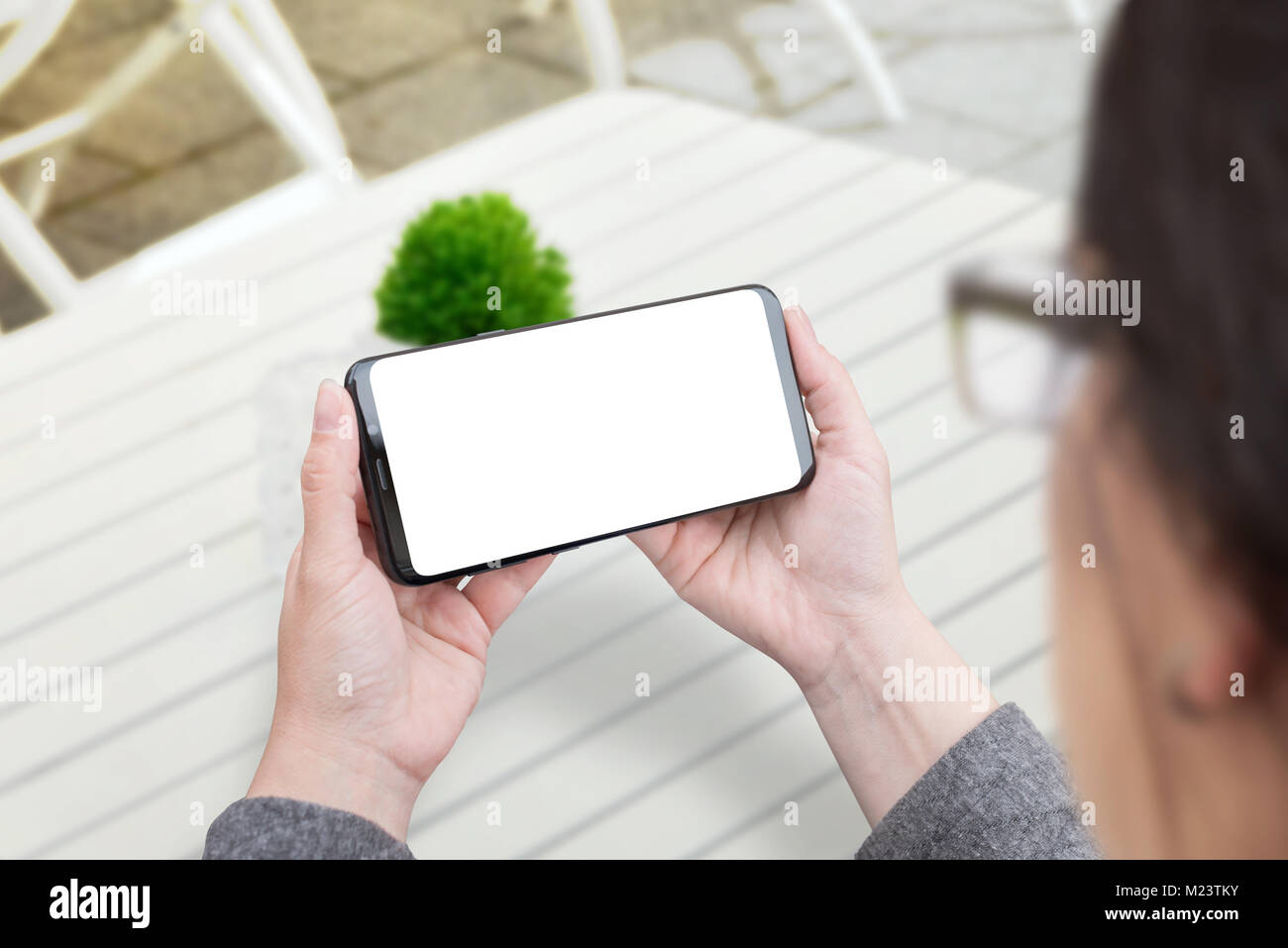 Woman holding smart phone in horizontal position. Isolated screen for mockup. White wooden desk and plant in background. Stock Photo