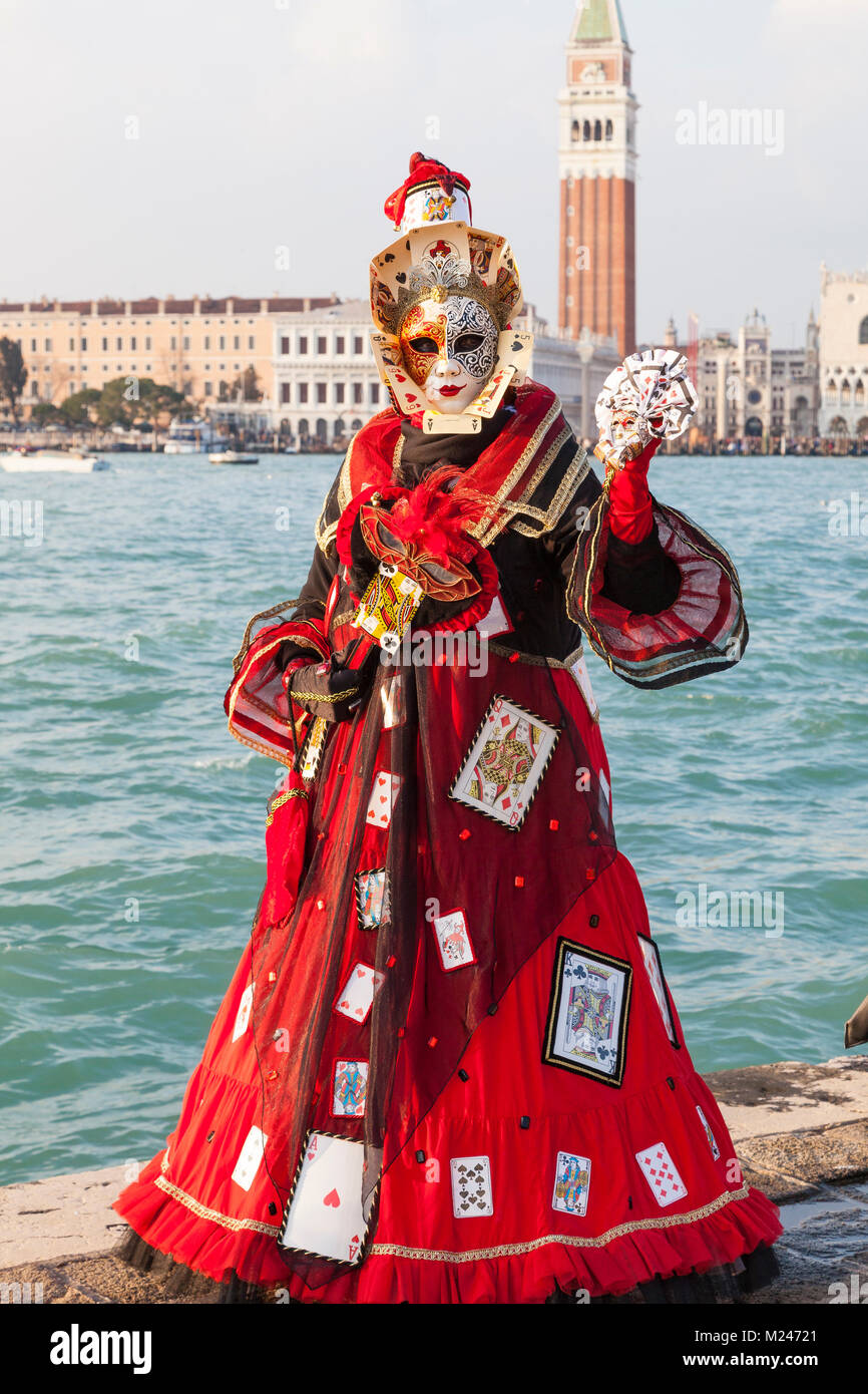 Venice, Veneto, Italy 4th February 2018. Woman wearing an unusual colourful red costume  and mask decorated with playing cards at San Giorgio Maggiore in late evening light with the lagoon and Campanile of St Marks Basilica behind her  at the Venice Carnival . Stock Photo