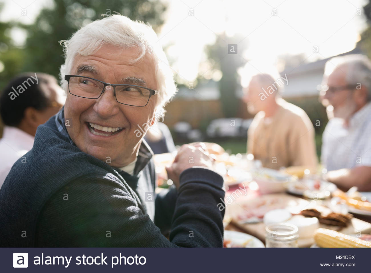 Smiling senior man looking over shoulder, enjoying garden party lunch at sunny patio table Stock Photo