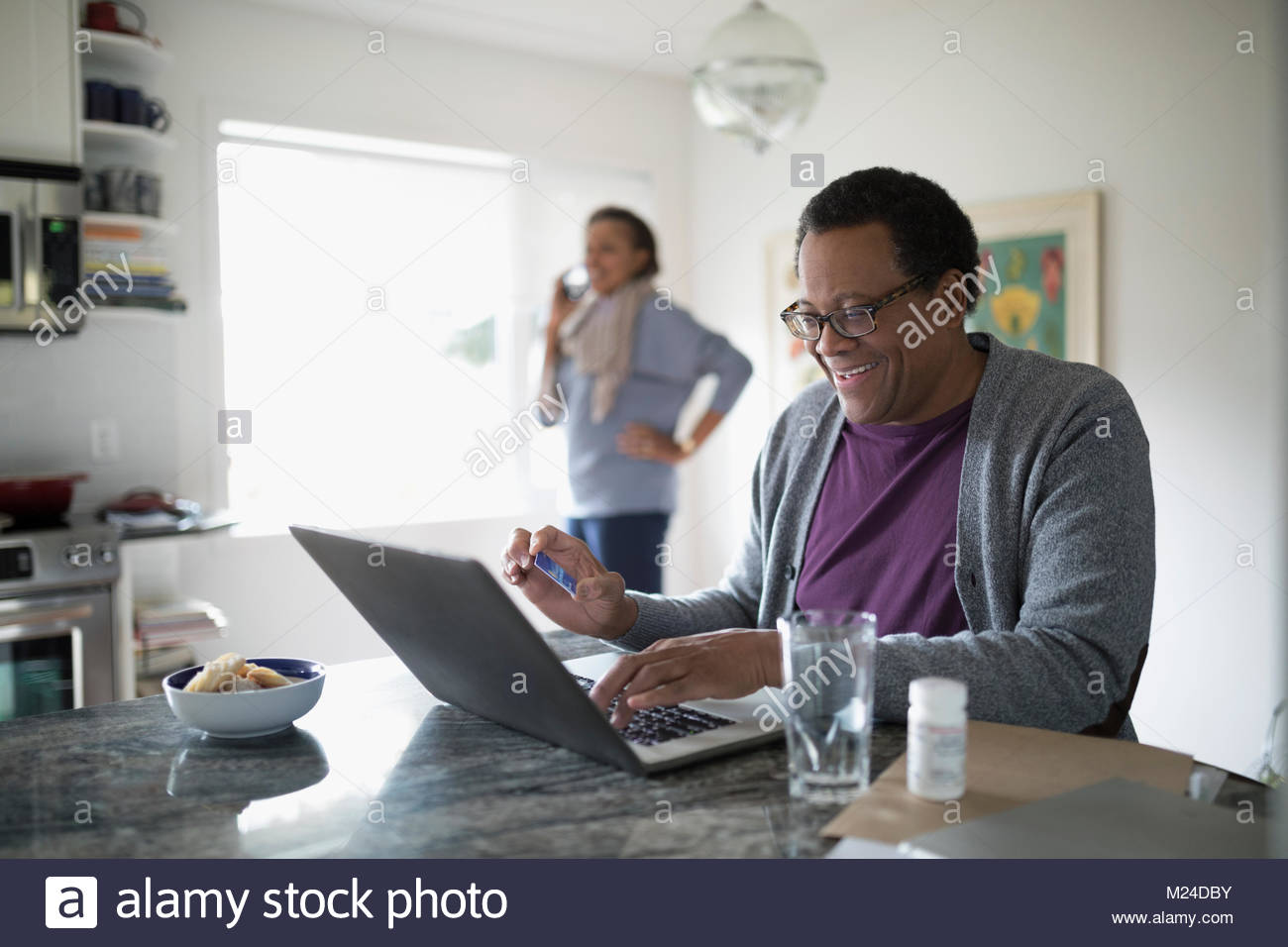 Smiling senior man with credit card paying bills online at laptop at kitchen counter Stock Photo