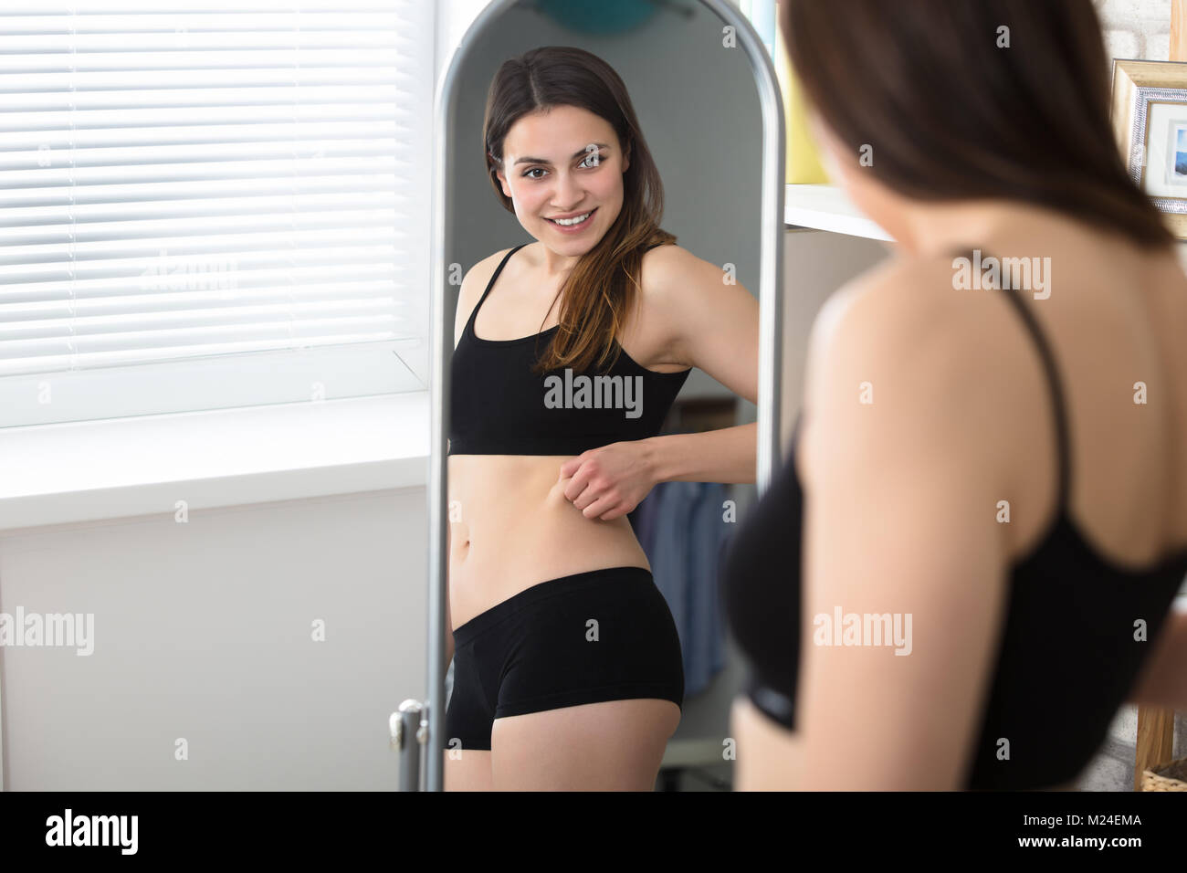 Young Woman Looking At Herself In The Mirror And Pinching Her Stomach Fat Stock Photo