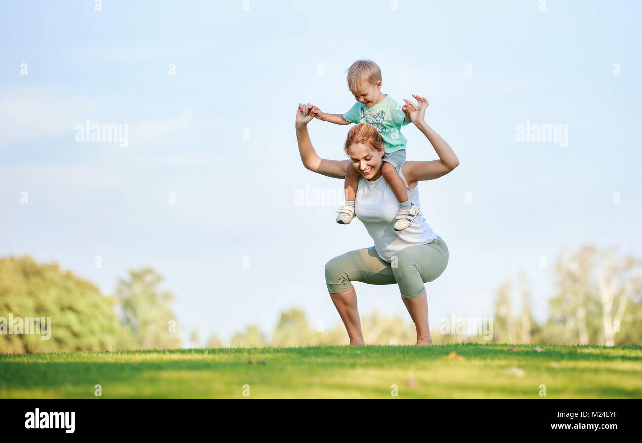 Young fitness woman working out outdoors while hoding son on shoulders Stock Photo