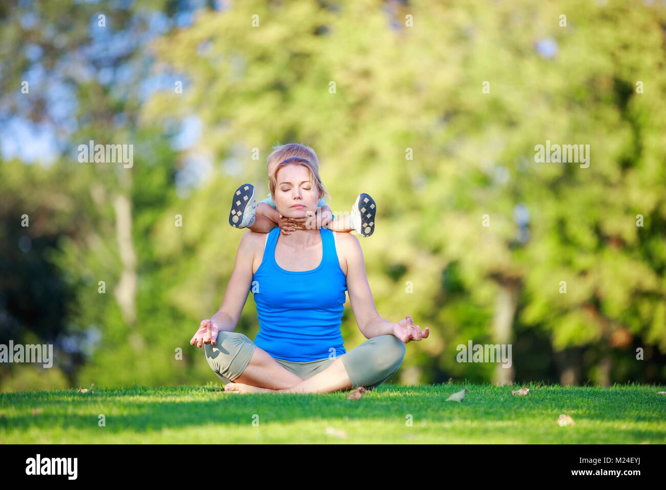 Young woman sitting in lotus position while holding little son on shoulders in summer park Stock Photo