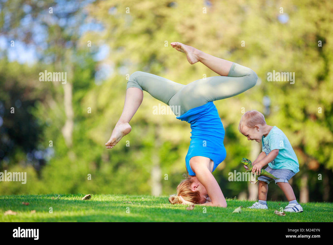 Young woman working out outdoors, standing on head, little son playing beside her Stock Photo