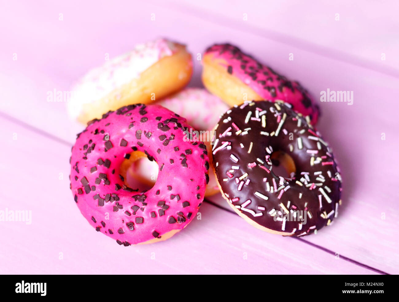 Delicious glazed donuts with sprinkles. Pink table and arrangement of chocolate donuts, various donuts, sweet food or unhealthy eating scene. Stock Photo