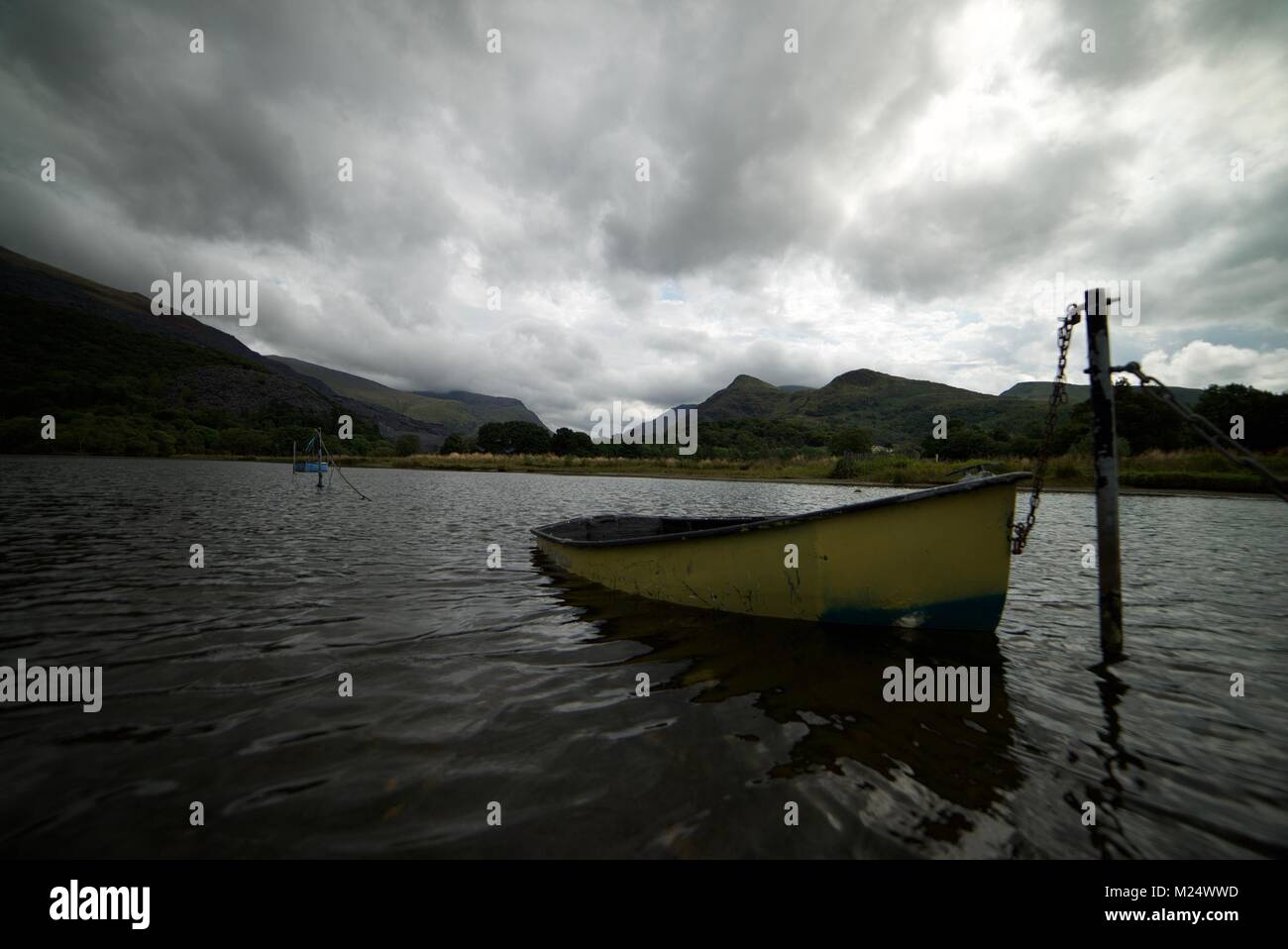 Yellow wooden paddle boat tied up on a lake in Snowdonia National Park, with a cloudy sky overhead surrounded by mountains Stock Photo