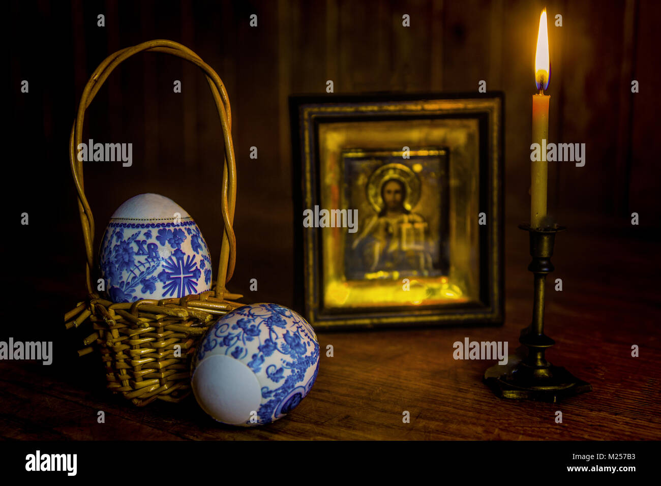 Easter eggs and a burning candle in a candlestick in front of an icon of Jesus Christ on a wooden table against a wall of wood. Stock Photo