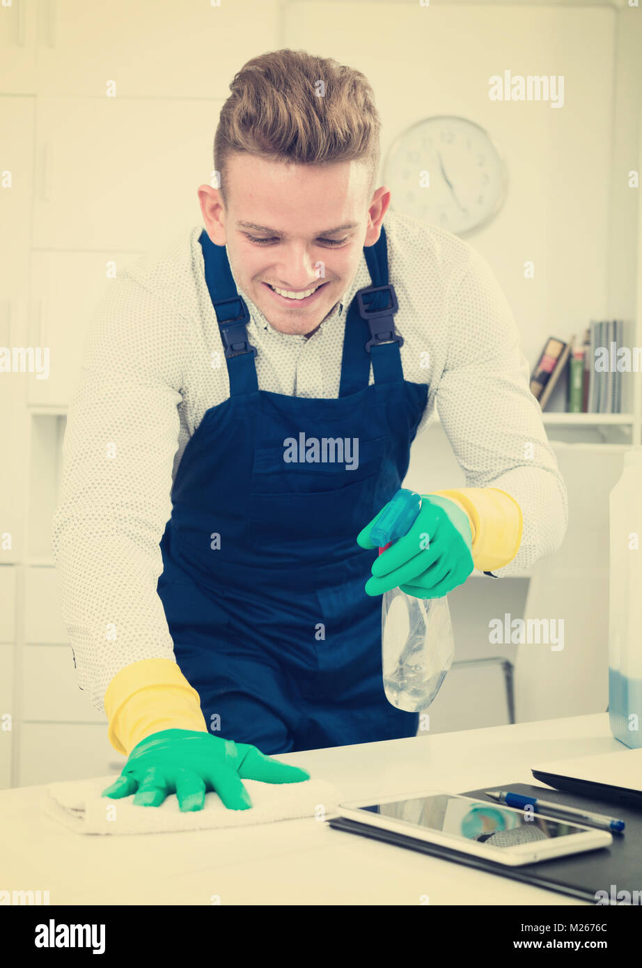Young male janitor dusting office desk and smiling indoors Stock Photo