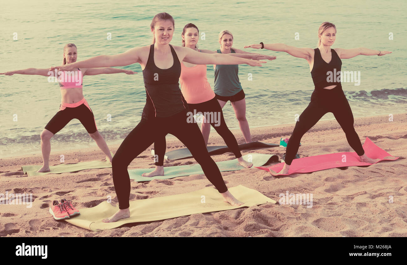 Smiling women exercising yoga asanas on sandy beach in sunny morning ...