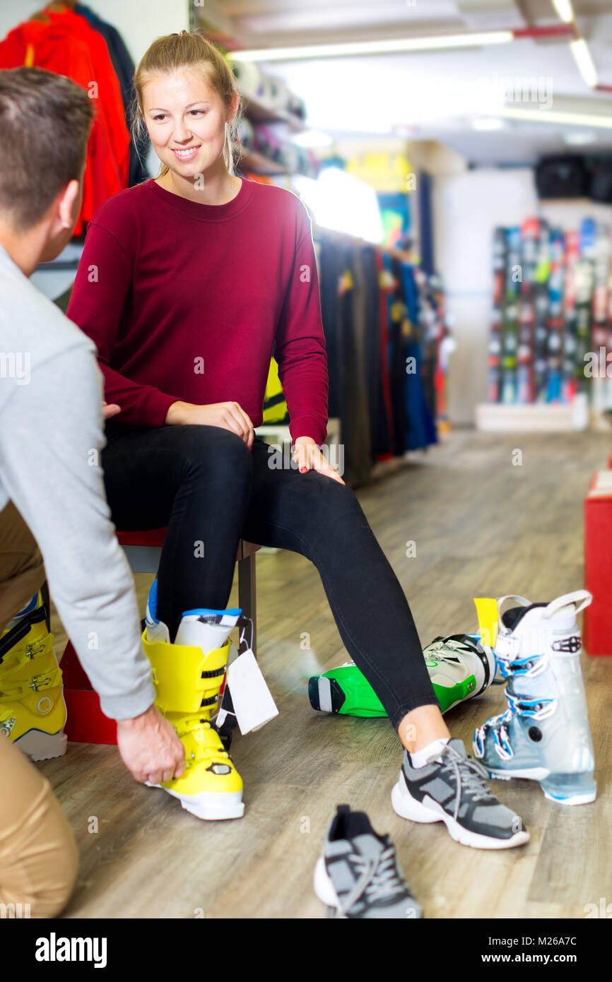 Glad shop assistant is helping woman to trying on ski boots in store