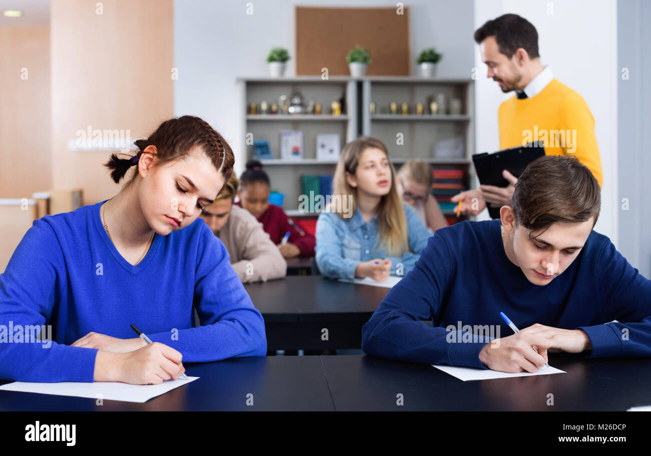 Positive  smiling teacher monitoring students’ work during examination test in class Stock Photo