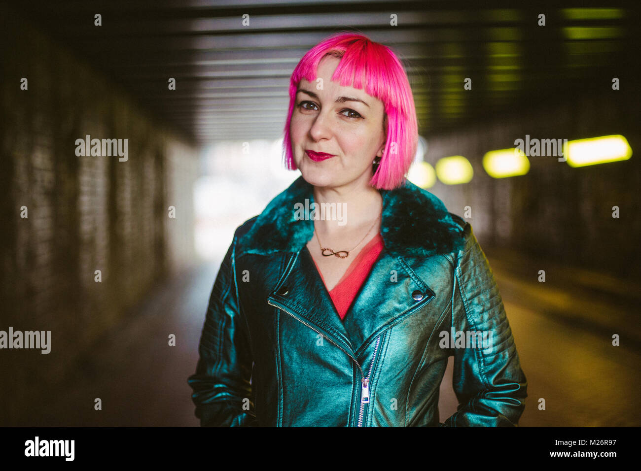 A woman with pink hair stands in an alley way looking bold. Stock Photo