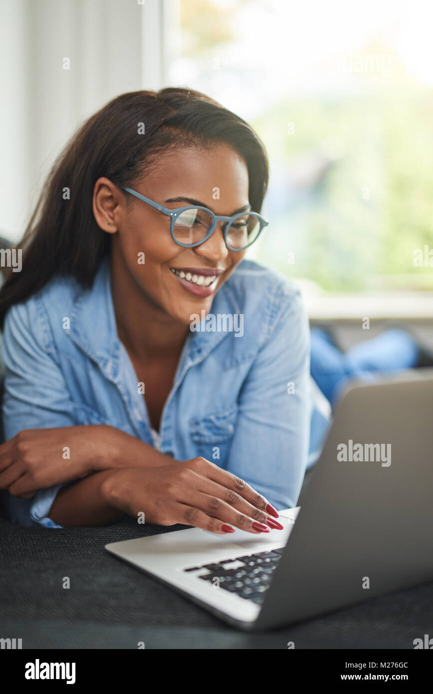 Smiling young African woman lying on a couch in her living room using a ...