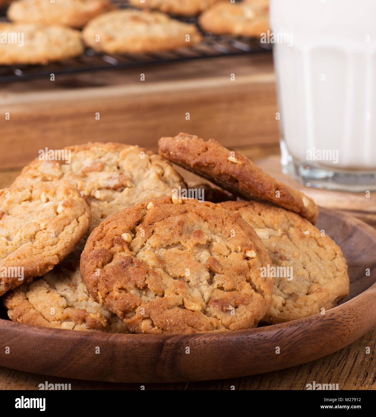 Closeup of peanut butter cookies on a wooden plate with cookies on a cooling rack and glass of milk in background Stock Photo