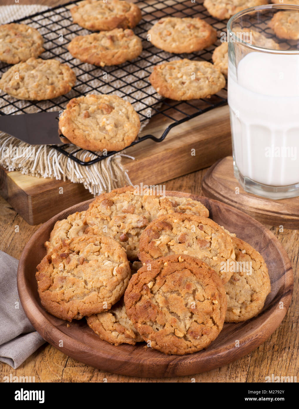 Peanut butter cookies on a wooden plate with cookies on a cooling rack and glass of milk in background Stock Photo