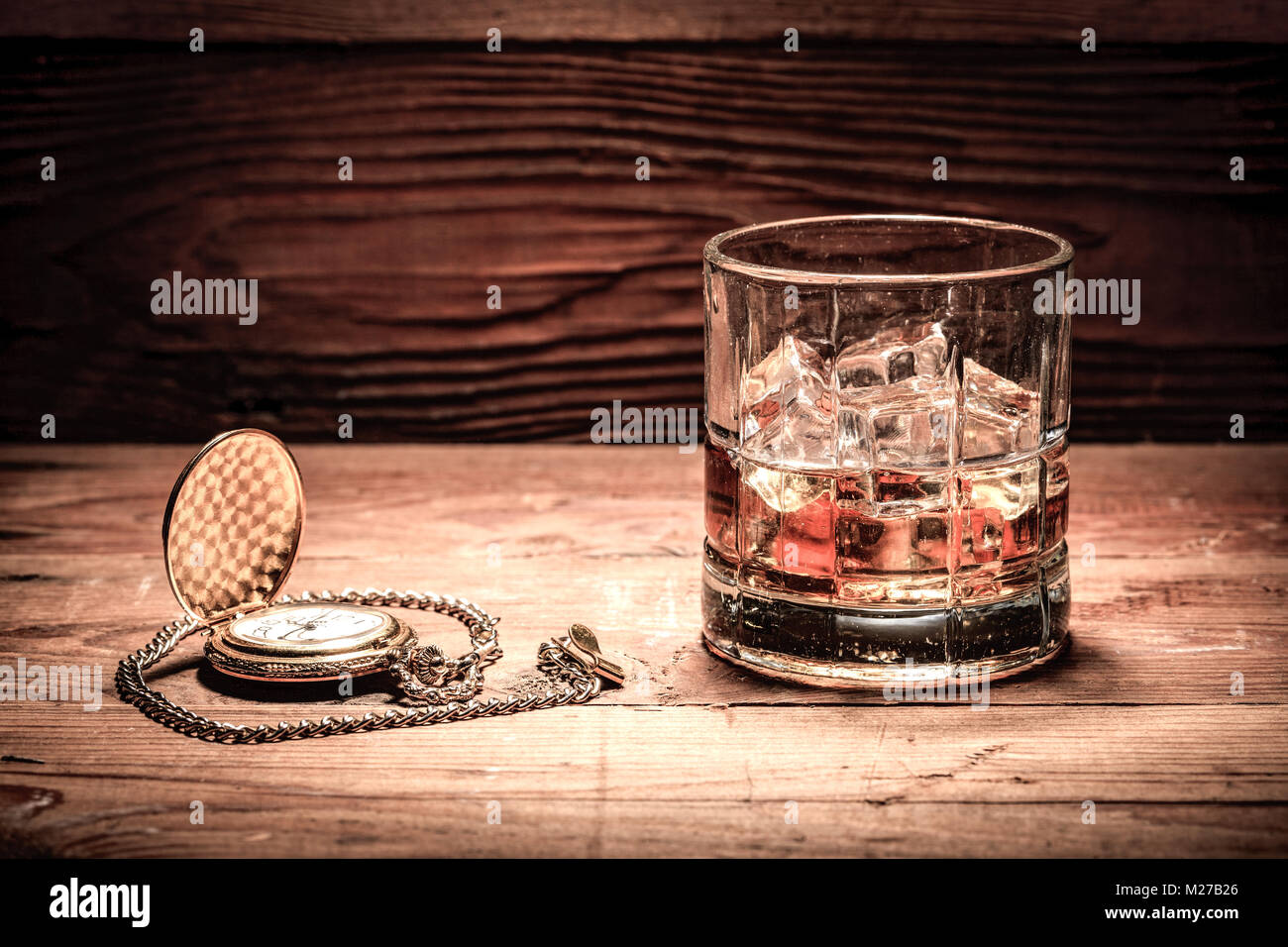 A retro style image of a glass of liquor with ice and a pocket watch on wood. Stock Photo