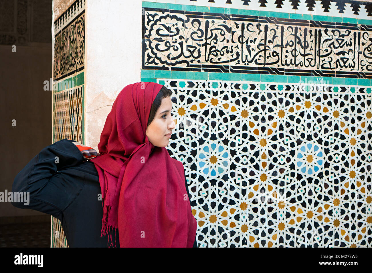 Arab woman in traditional clothing with red hijab in front of a wall with text from Koran Stock Photo