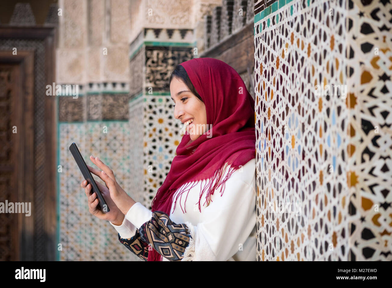 Smiling Arab woman in traditional clothing with red headscarf on her head working on tablet Stock Photo