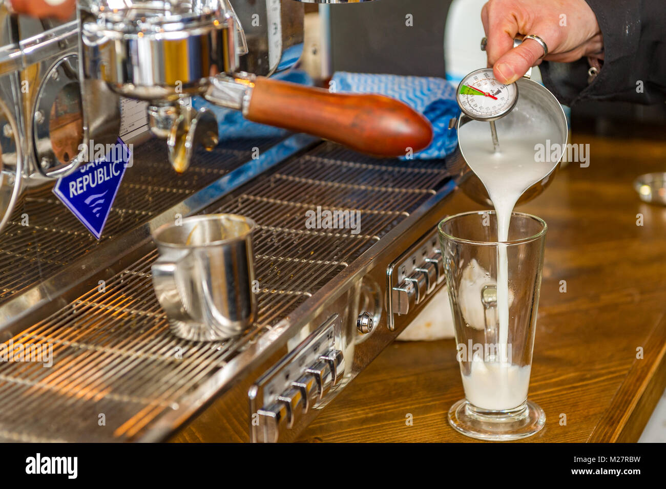 Barista pouring hot milk into a latte glass with milk thermometer to make a latte. Stock Photo