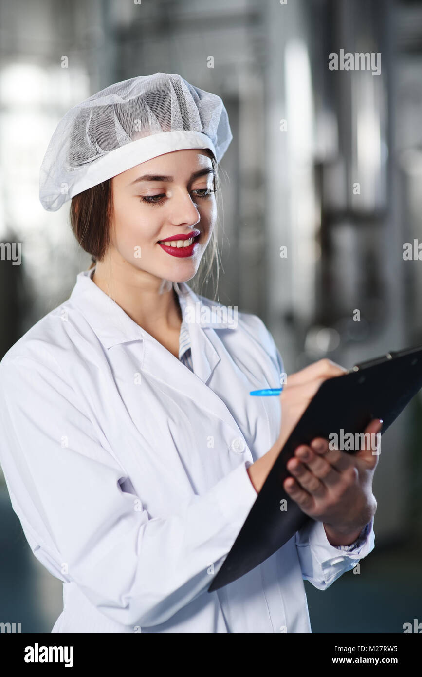 beautiful young girl in white working clothes is making a note on paper against the background of factory equipment Stock Photo